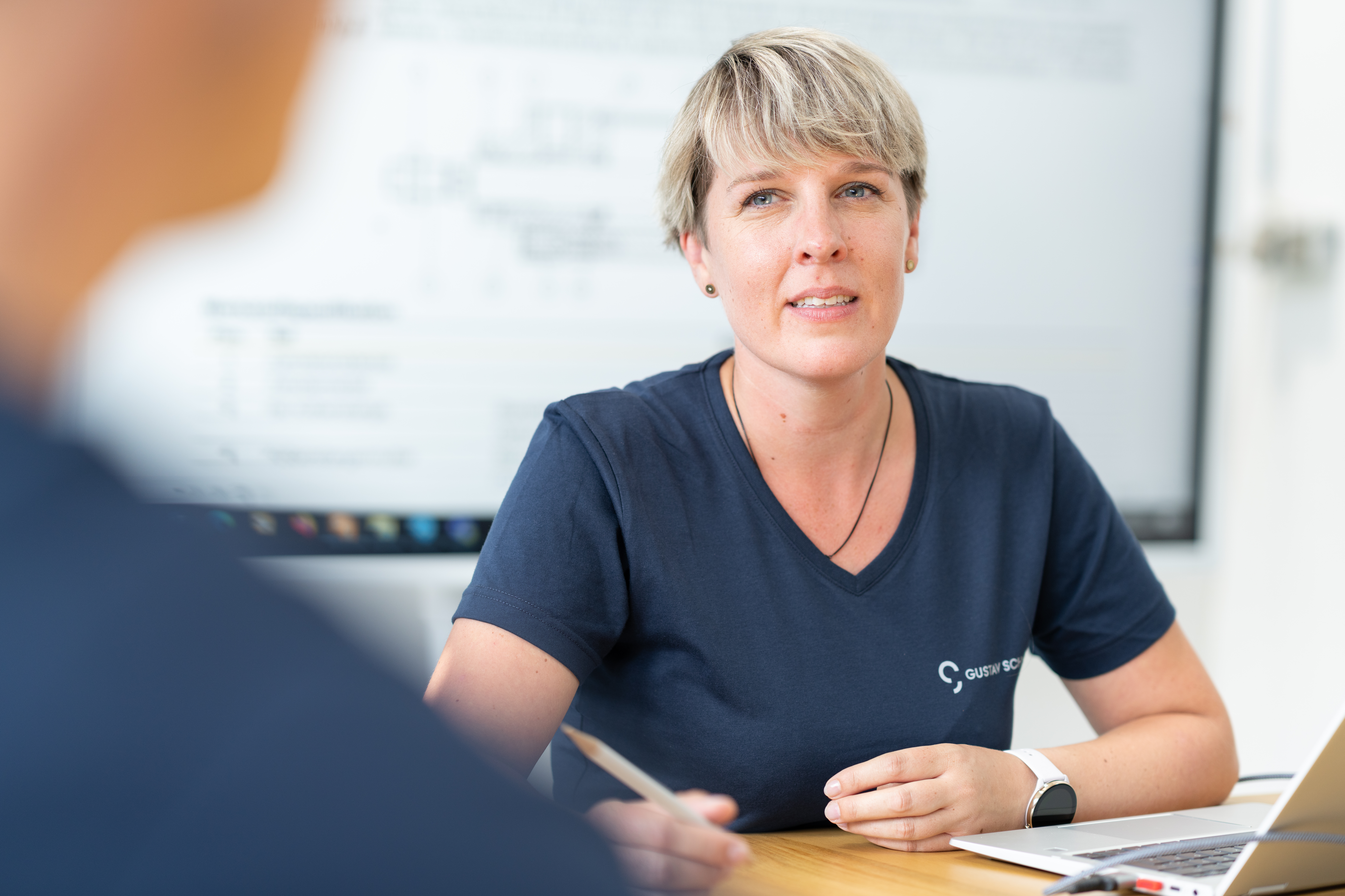 Frau mit blonden Haaren und blauem Gustav-Schmidt-Shirt schaut andere Person (nur verschwommen sichtbar) mit lächelndem Gesicht an, im Hintergrund digitale Präsentationstafel zu sehen, am Tisch vor ihr ein Laptop und hat Stift in der Hand